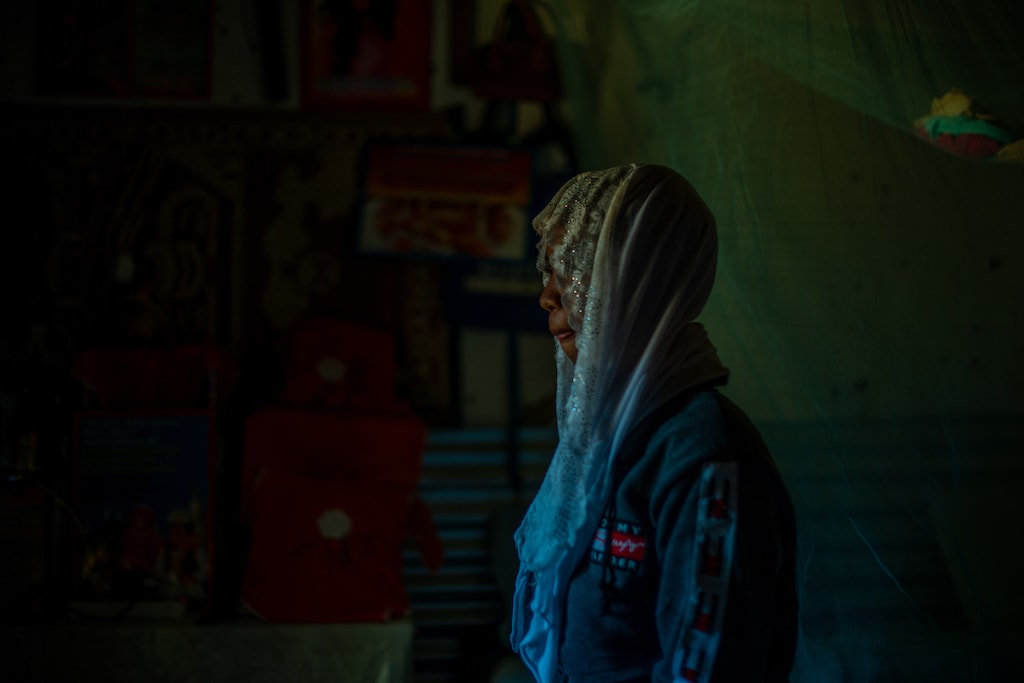 Maria is standing in a dark room. She is wearing a dark blue dress and a head covering.